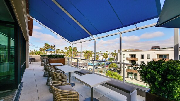 An outdoor patio with tables and chairs, a blue awning, and a view of the cityscape. - Steps to the Sand! Luxury Oceanside Pier Condo (Oceanside)