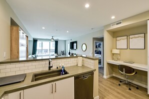 A modern kitchen with a dishwasher, a sink, and a countertop. There is a dining area with a table and chairs visible in the background. - Steps to the Sand! Luxury Oceanside Pier Condo (Oceanside)