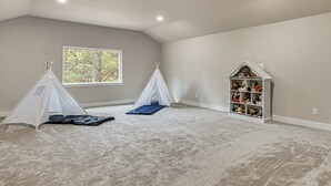 Ein geräumiges Zimmer mit hoher Decke, Teppichboden, einem Fenster mit Blick auf Bäume, zwei weiße Spielzelte und ein weißes Bücherregal voller Stofftiere.