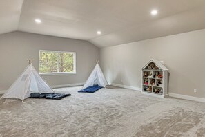 Ein geräumiges Zimmer mit hoher Decke, Teppichboden, einem Fenster mit Blick auf Bäume, zwei weiße Spielzelte und ein weißes Bücherregal voller Stofftiere.