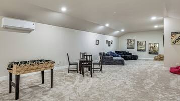 Um quarto com mesa de pebolim, mesa de jantar com cadeiras, sofá e vários cartazes emoldurados na parede.
