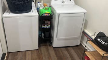 Uma lavanderia com máquina de lavar, secadora, cesto de roupa e prateleira com produtos de limpeza.