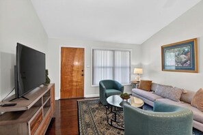 A living room with a television, a wooden door, a window with blinds, a sofa, a chair, a small table, and a lamp. - Golf & Chill! Family-friendly Augusta Home (Augusta)