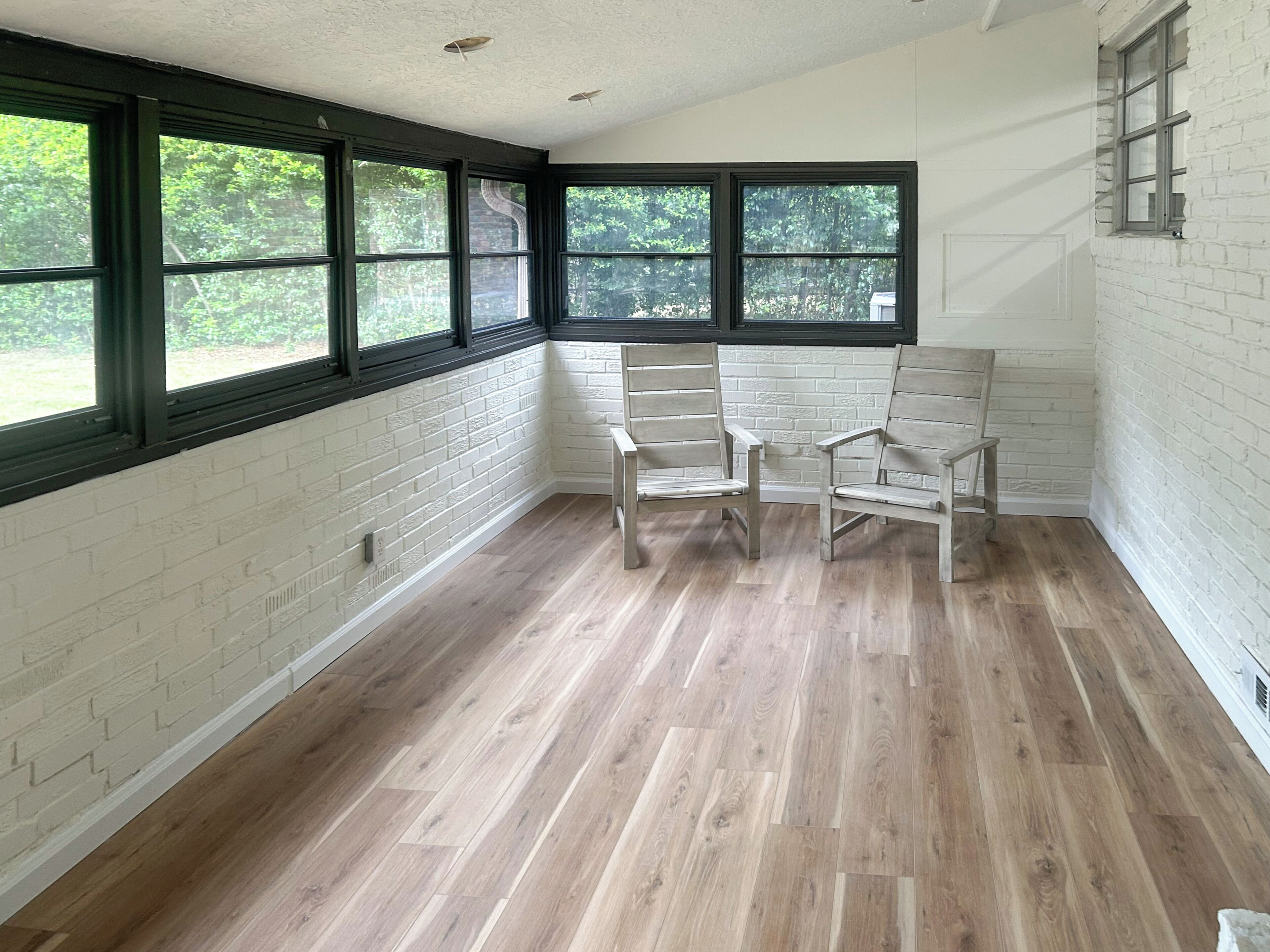 A room with white brick walls, wooden flooring, and black-framed windows. There are two wooden chairs and a white bench.
