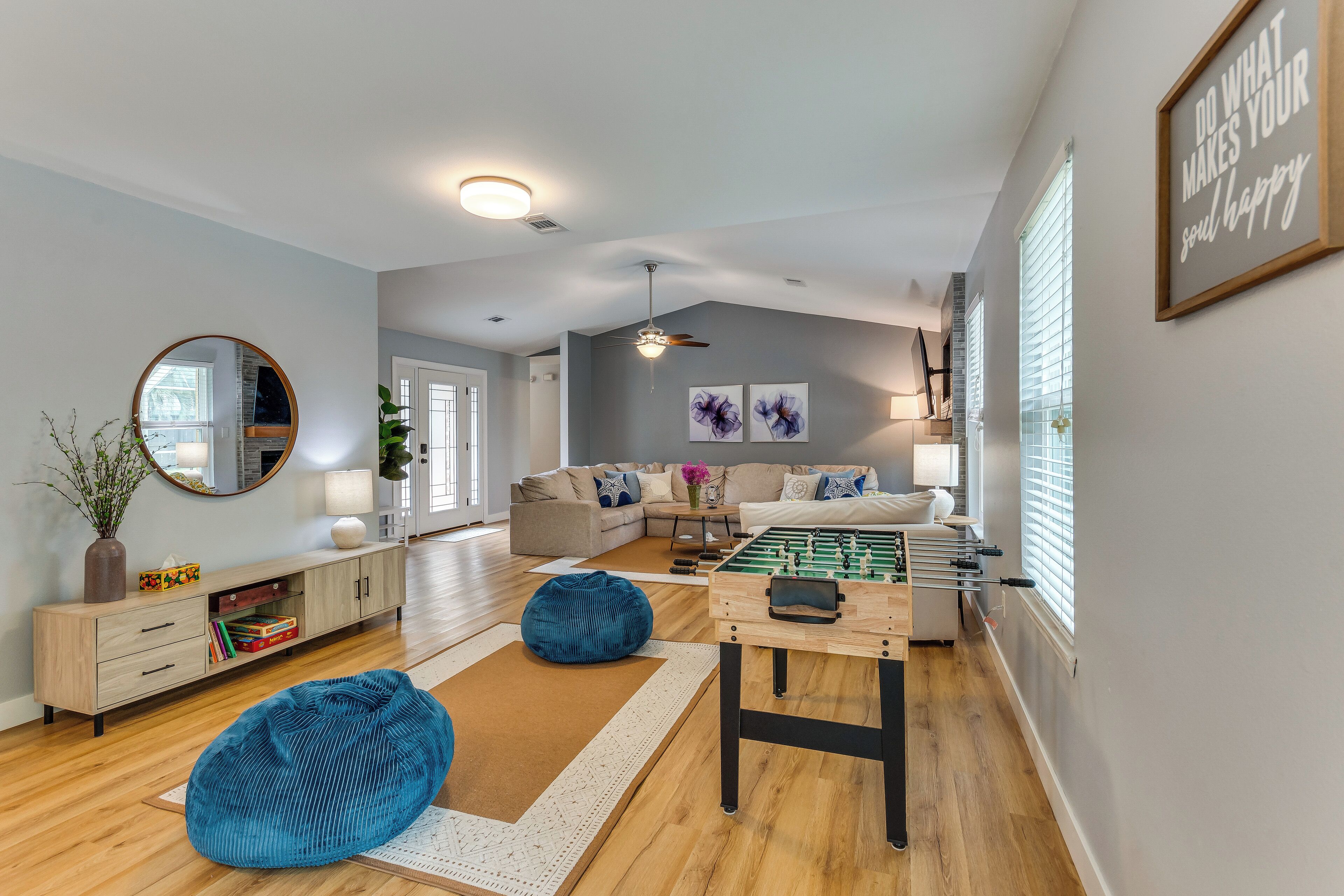 A modern living room with a sofa, coffee table, and foosball table. There is a mirror, a plant, and a framed quote on the wall.