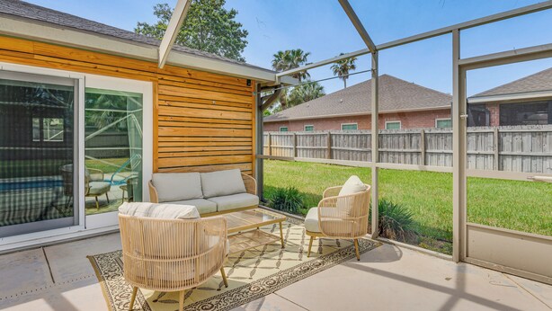 A covered patio area with wicker furniture, a glass door leading outside, and a view of a backyard with a wooden fence. - Pool Days! Family Getaway Near Pensacola Beach (Gulf Breeze)