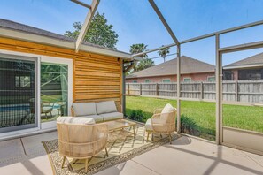 Ein überdachter Patio-Bereich mit Korbmöbeln, einer Glastür, die nach draußen führt, und Blick auf einen Garten mit Holzzaun.