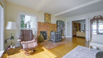 A spacious living room with a large window, a fireplace, a rocking chair, and a kitchen area visible through an open door.