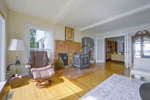 A spacious living room with a large window, a fireplace, a rocking chair, and a kitchen area visible through an open door. - Charming Waterfront Home With Mt Rainier View (Olympia)