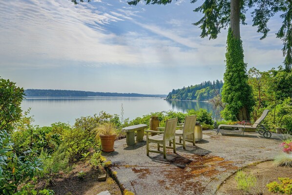 A lakeside seating area with chairs and a bench, surrounded by greenery and overlooking a calm lake. - Charming Waterfront Home With Mt Rainier View (Olympia)