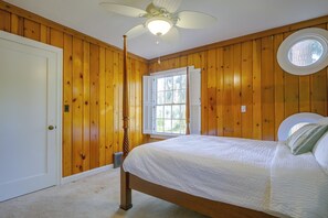 A bedroom with wooden walls, a bed with a white bedspread, a ceiling fan, and a window with shutters. - Charming Waterfront Home With Mt Rainier View (Olympia)