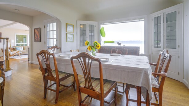 Een eetkamer met een ronde tafel gedekt voor zes personen, houten stoelen, een vaas met zonnebloemen en uitzicht door een raam.