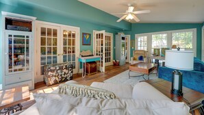 A room with teal walls, a ceiling fan, a white sofa, a desk with a lamp, and a window with a view of trees. - Group Getaway w/ Private Yard in Marietta (Marietta)