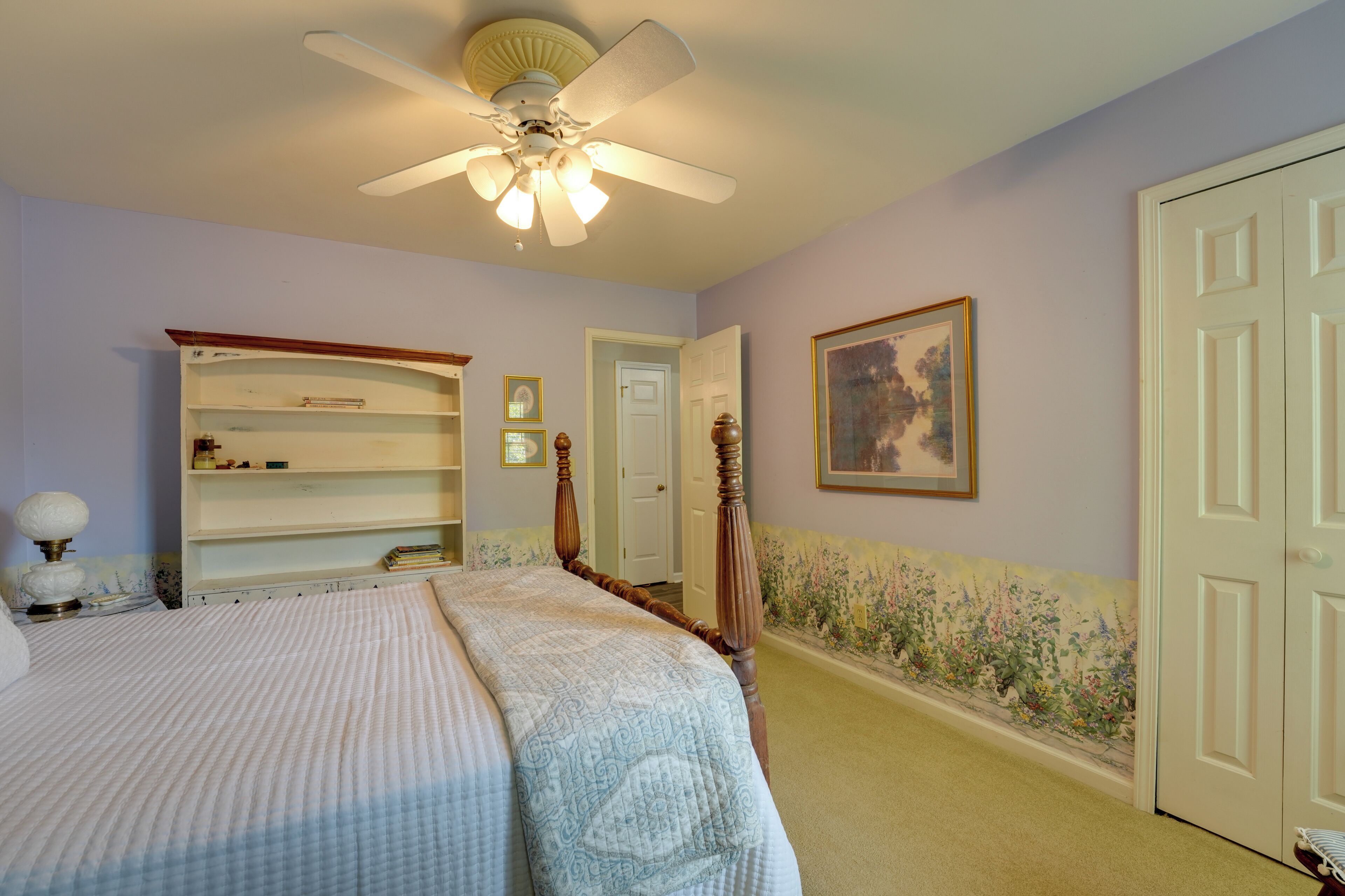 A bedroom with a bed, a ceiling fan, a bookshelf, a door, and a wall with a floral wallpaper border.
