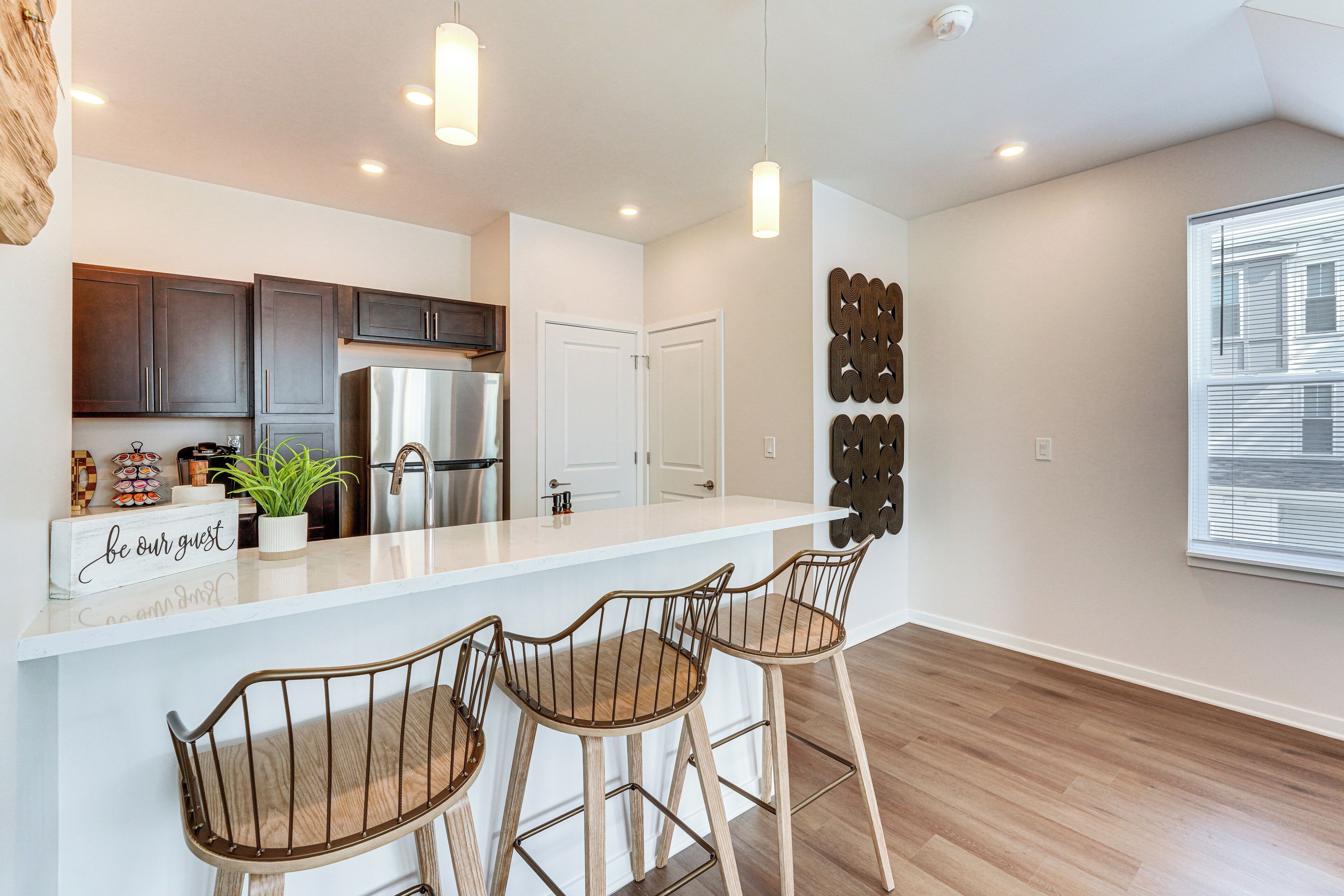 A modern kitchen with a stainless steel refrigerator, dark wooden cabinets, and a white countertop. There are two bar stools with metal frames and wooden seats.
