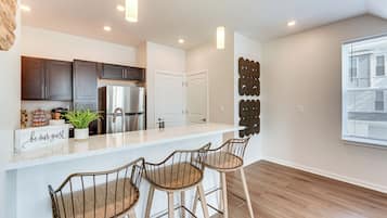 A modern kitchen with a stainless steel refrigerator, dark wooden cabinets, and a white countertop. There are two bar stools with metal frames and wooden seats.