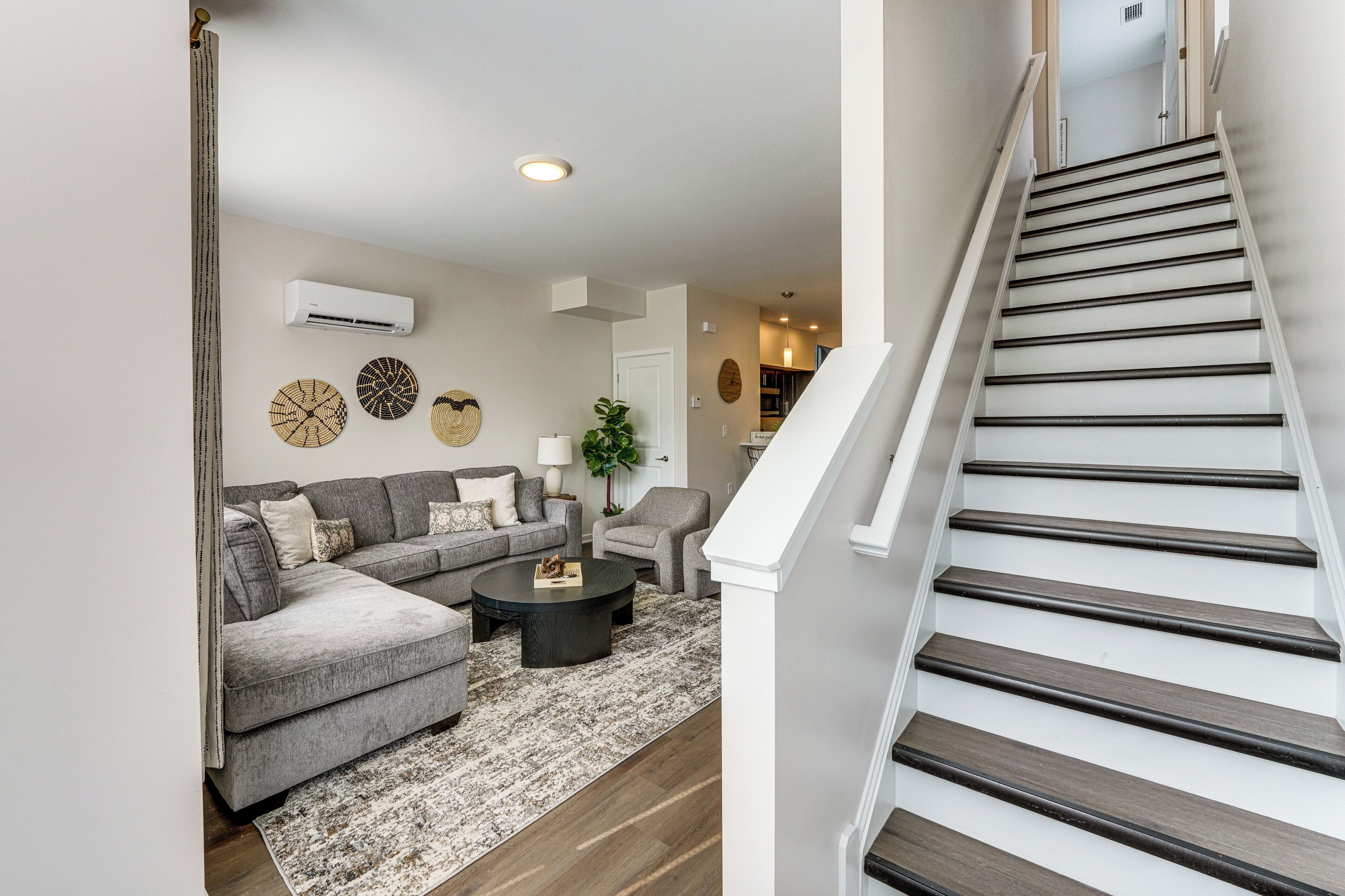 A modern living room with a grey sectional sofa, a round coffee table, and a patterned rug. A staircase with dark wooden steps and white railings leads to an upper level.