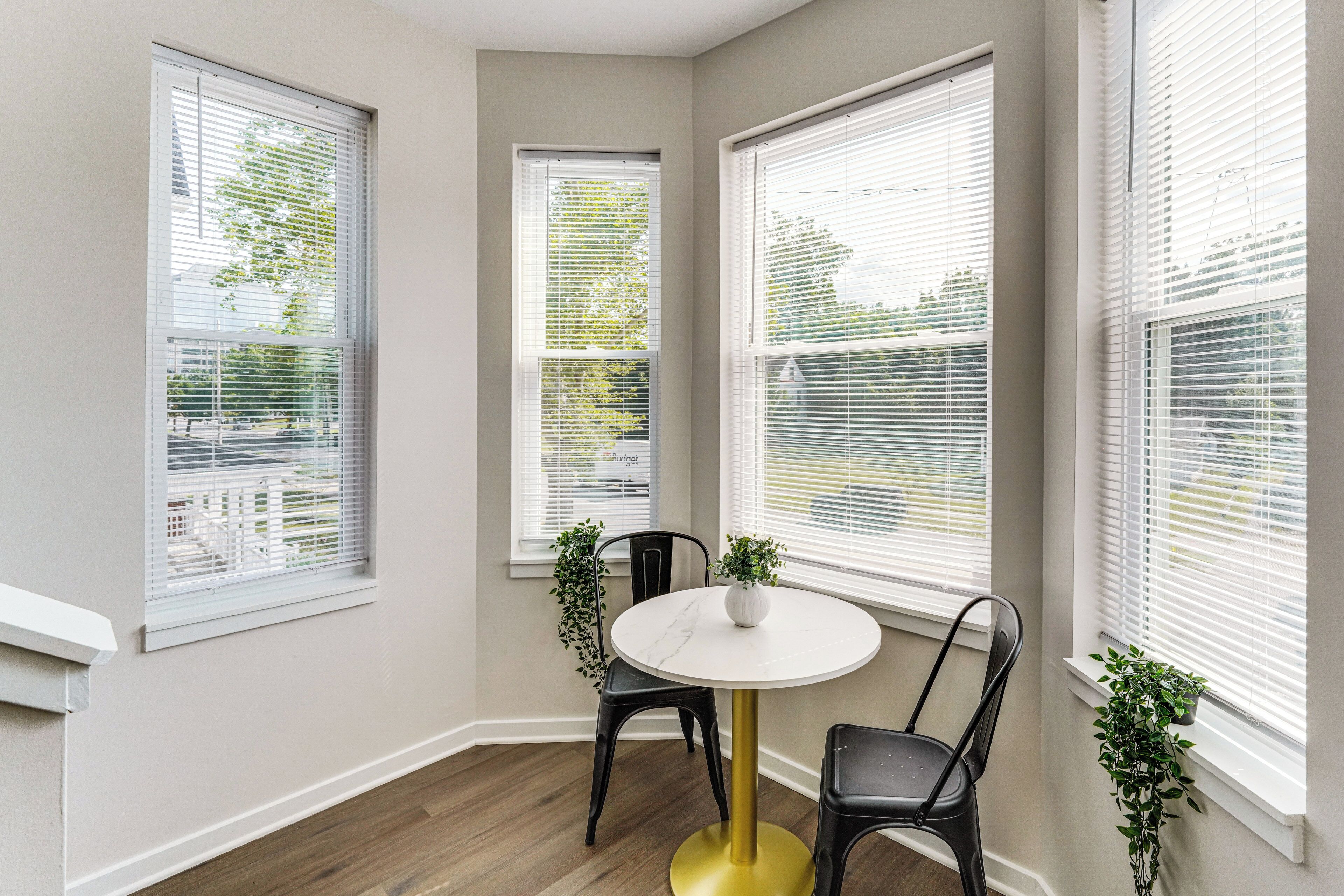 A corner room with a round table, two chairs, and three windows with blinds.