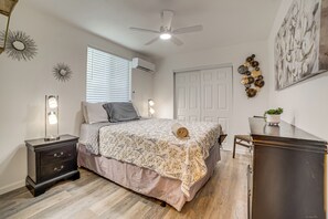 Ein Schlafzimmer mit Bett, Nachttischen, Schreibtisch, Stuhl, Deckenventilator, Fenster mit Jalousien, Klimaanlage und Wanddekorationen.
