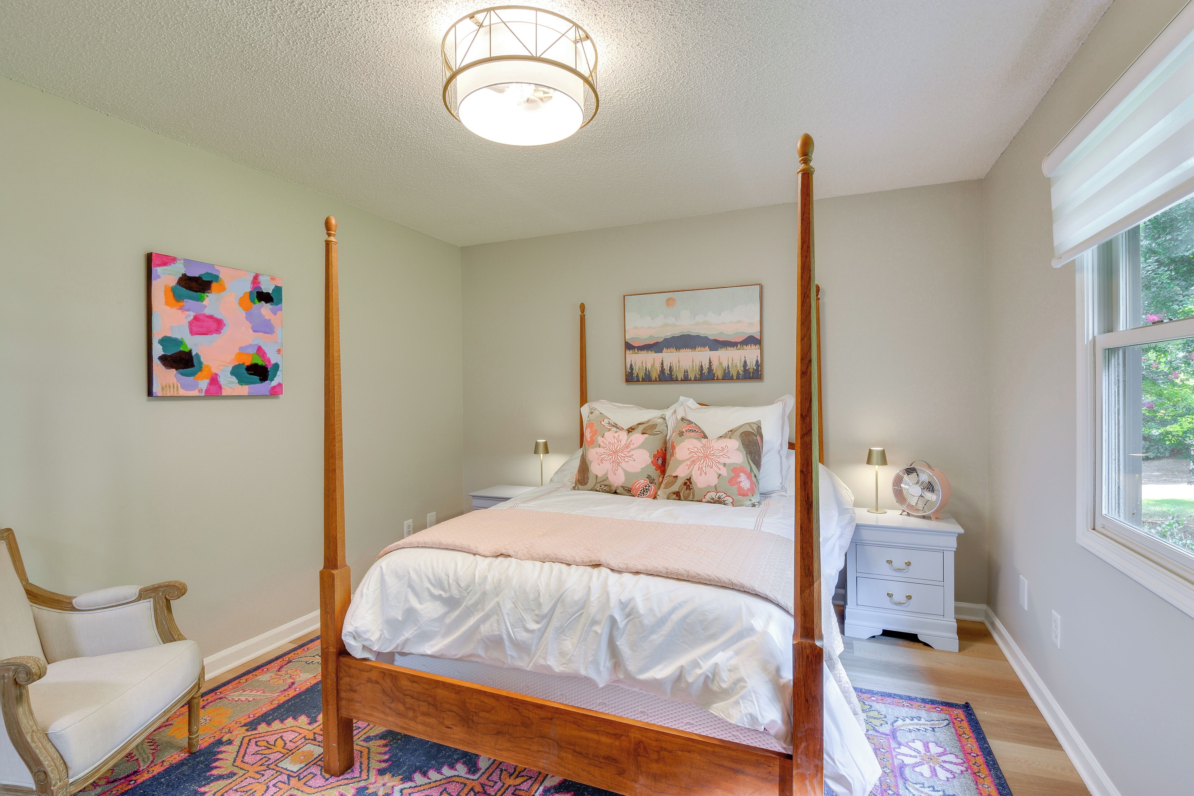 A four-poster bed with a white bedspread and pillows, a nightstand with a lamp and a fan, a chair, and a colorful rug.