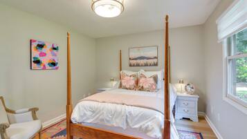 A four-poster bed with a white bedspread and pillows, a nightstand with a lamp and a fan, a chair, and a colorful rug.