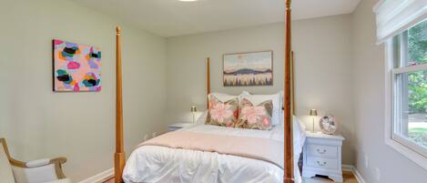 A four-poster bed with a white bedspread and pillows, a nightstand with a lamp and a fan, a chair, and a colorful rug.