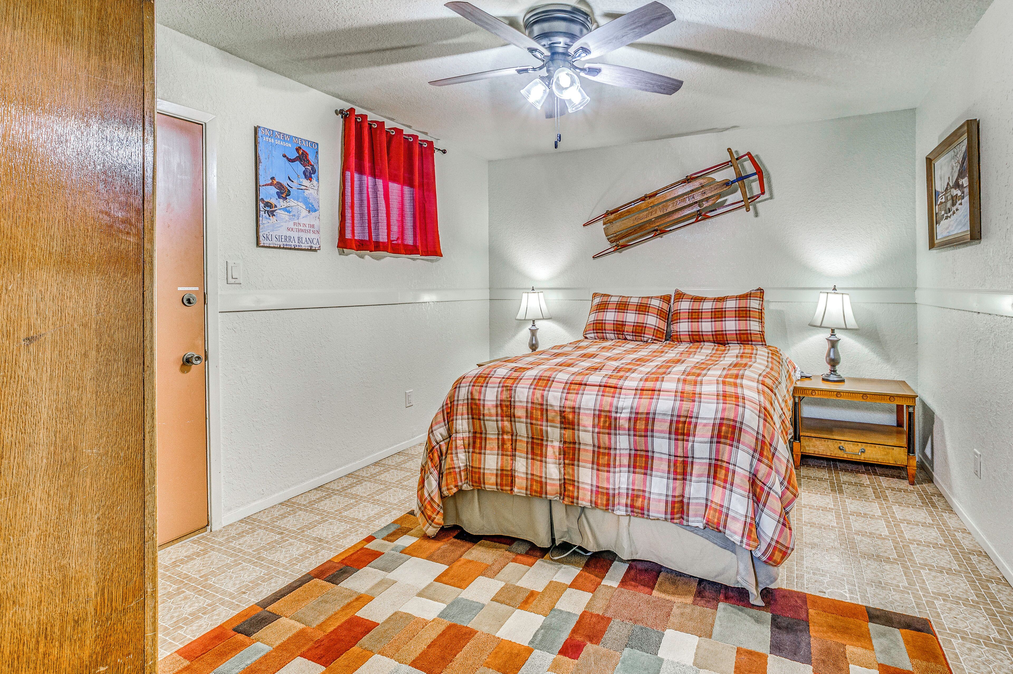 A bedroom with a bed, two bedside lamps, a ceiling fan, a red curtain, a poster on the wall, and a wooden sled decoration.