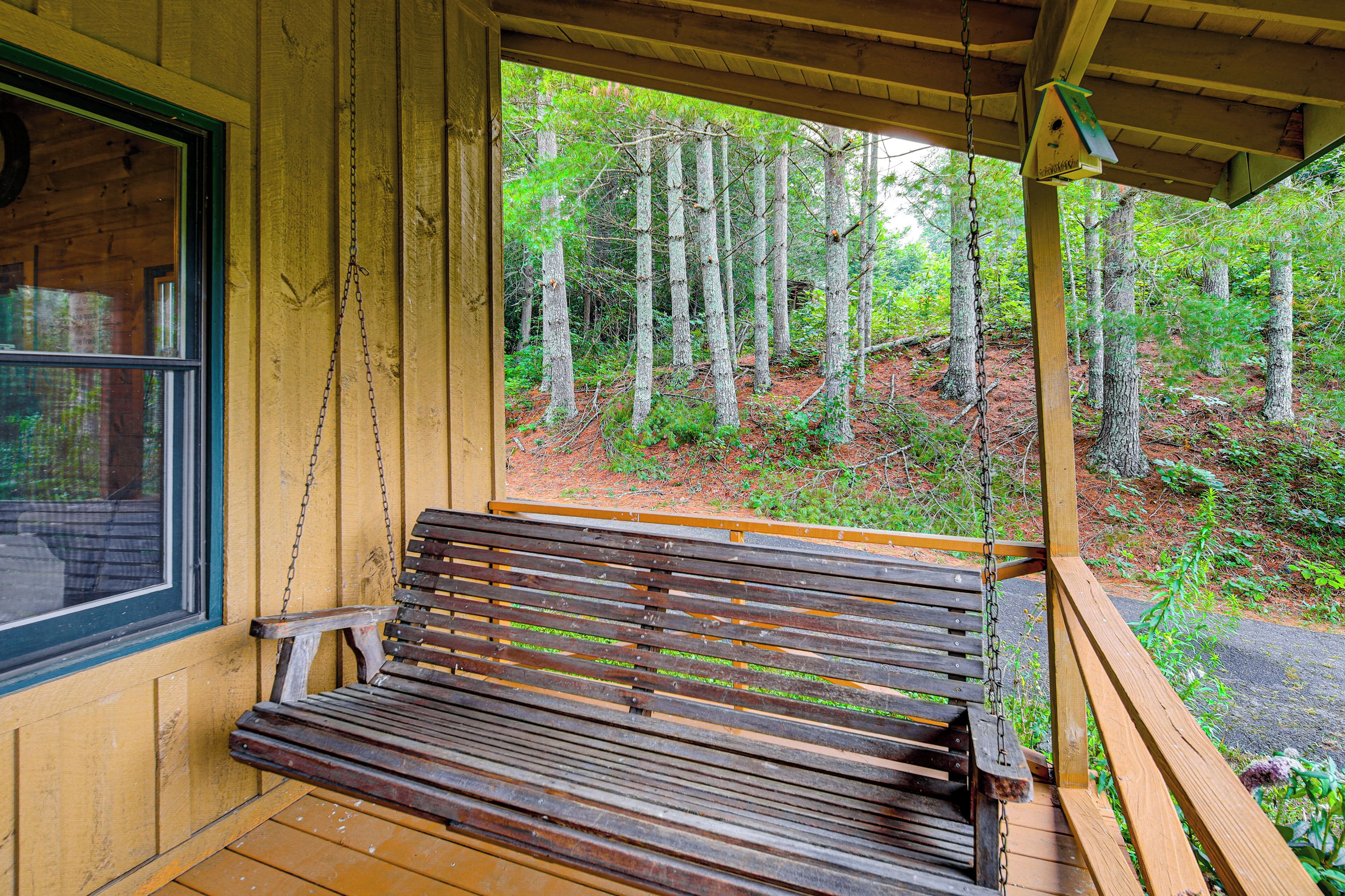 A wooden bench with a chain link backrest, positioned on a porch with a view of a forested area.