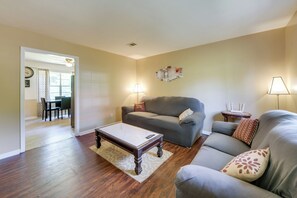A living room with a grey sofa, a coffee table, and two lamps. There is a dining area visible through an open door.