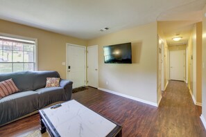 A living room with a gray sofa, a coffee table, a flat-screen TV mounted on the wall, hardwood flooring, and a door leading to another room.