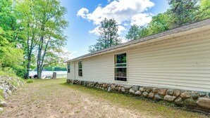 A single-story house with a stone foundation, a dirt yard, and a view of a lake. - Waterfront Escape on Bass Lake: Paddle, Fish, Swim (Irons)