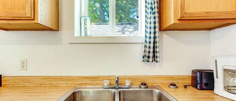 A kitchen with a double stainless steel sink, wooden cabinets, a window with a view of trees, and a microwave.