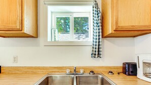 A kitchen with a double stainless steel sink, wooden cabinets, a window with a view of trees, and a microwave. - Waterfront Escape on Bass Lake: Paddle, Fish, Swim (Irons)