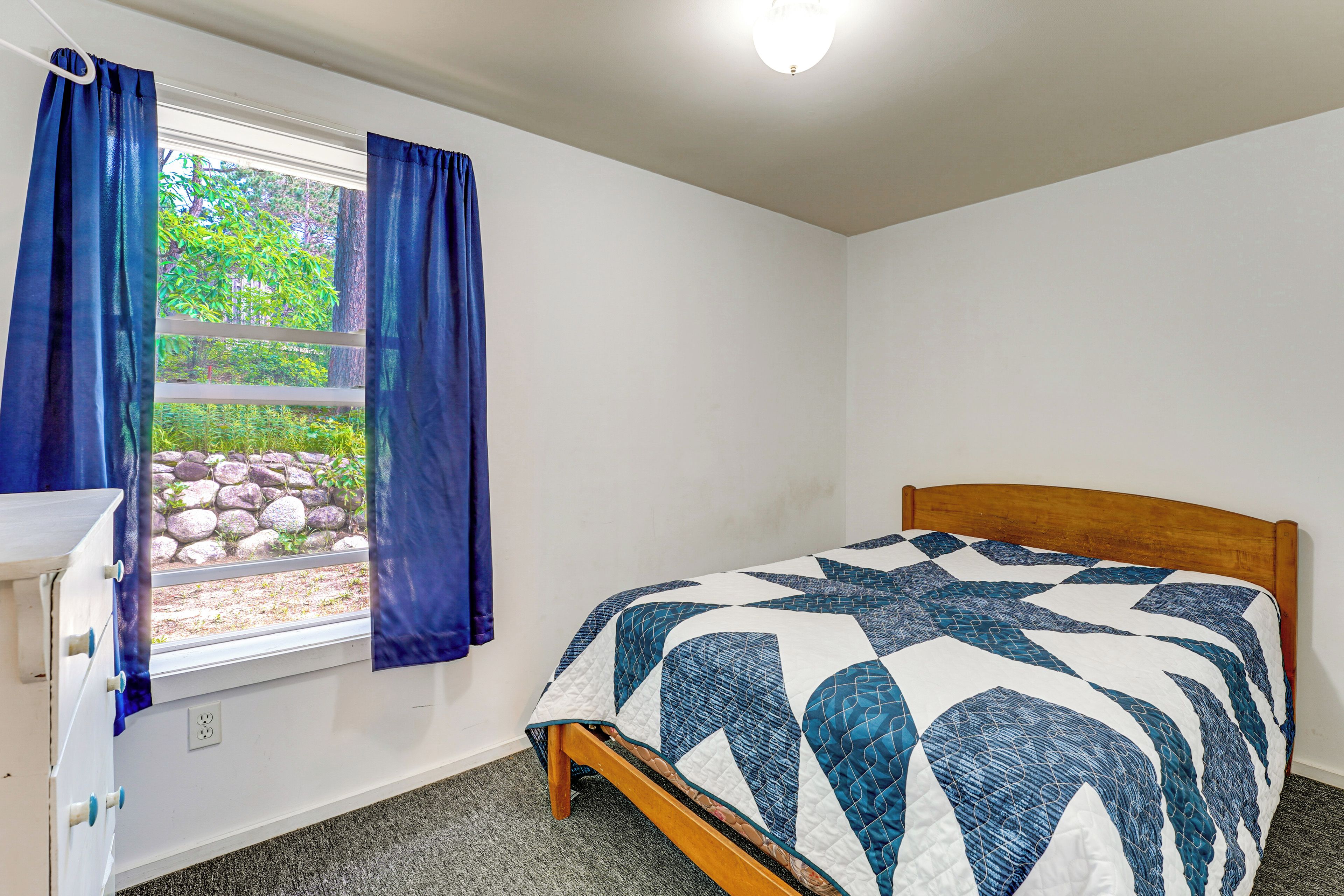 A bedroom with a bed, a window with curtains, a wooden headboard, and a white dresser.