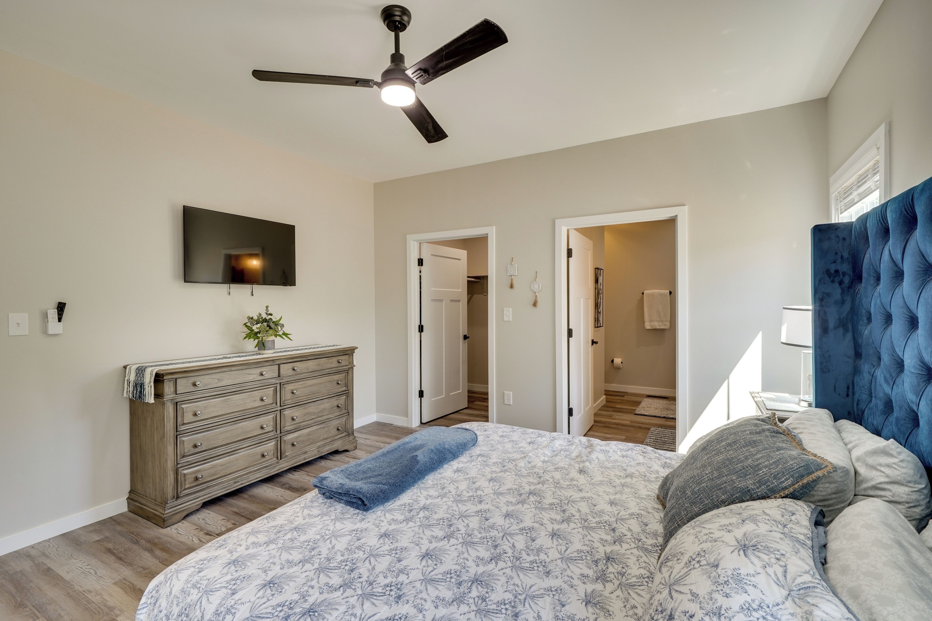 A bedroom with a bed, dresser, ceiling fan, TV, and a doorway leading to another room.