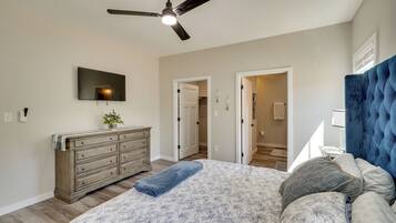 A bedroom with a bed, dresser, ceiling fan, TV, and a doorway leading to another room.