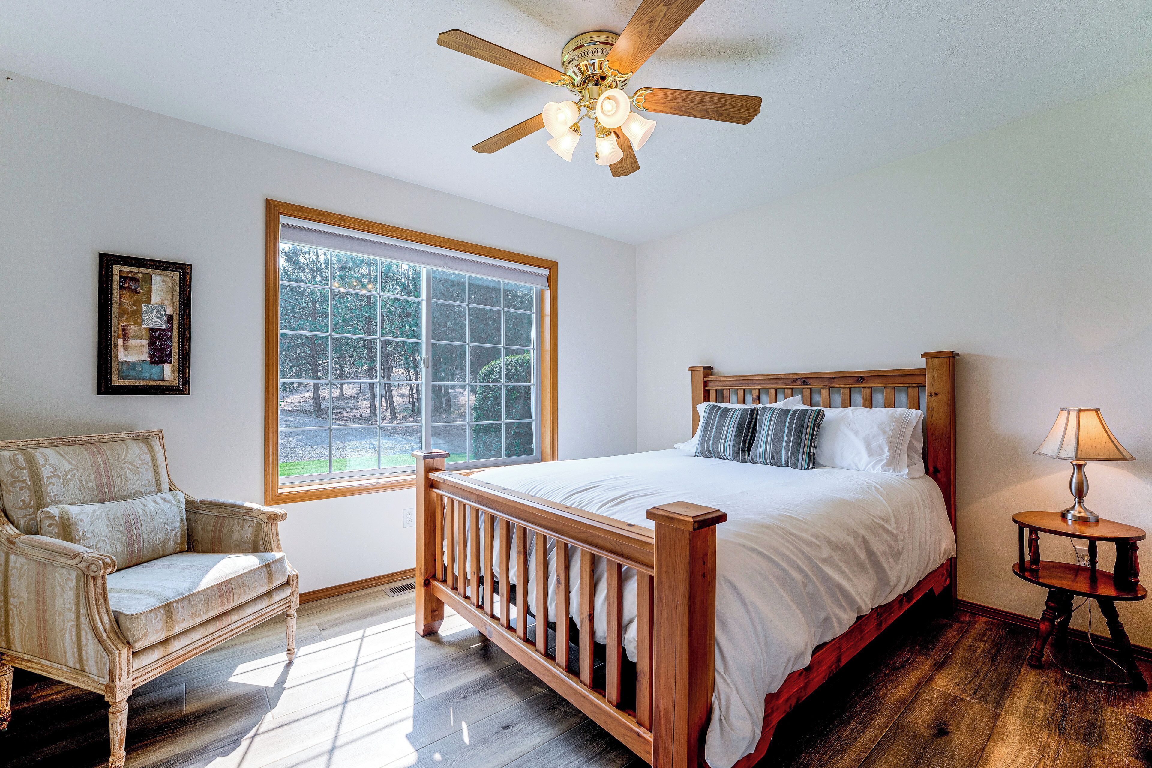 A bedroom with a wooden bed, a chair, a bedside table with a lamp, a ceiling fan, and a window with a view of trees.