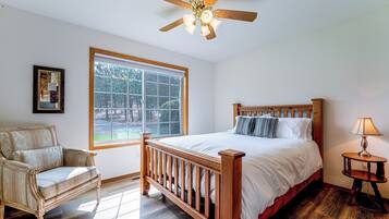 A bedroom with a wooden bed, a chair, a bedside table with a lamp, a ceiling fan, and a window with a view of trees.