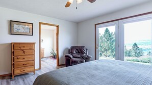 A bedroom with a large bed, a wooden dresser, a leather chair, and a door leading to a balcony with a view of trees. - Lake Roosevelt Retreat Lake: Mtn Views, Game Room (Davenport)