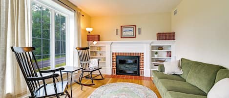 A living room with a fireplace, a green sofa, a rocking chair, and a view of the outdoors through large windows.