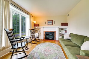 A living room with a fireplace, a green sofa, a rocking chair, and a view of the outdoors through large windows. - Farmhouse W/creek & Fireplace Near Gettysburg! (Biglerville)