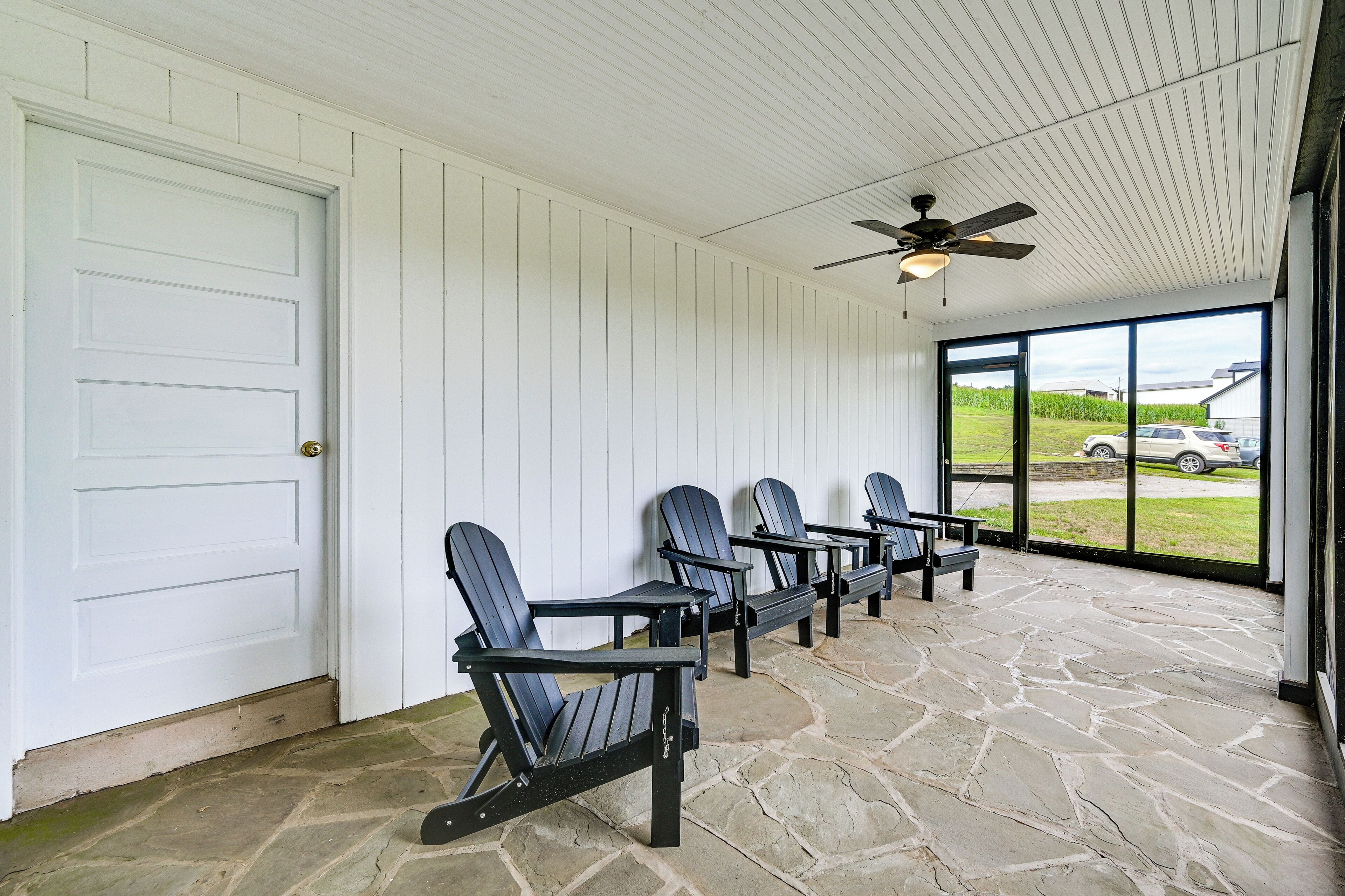 A covered porch with four Adirondack chairs, a ceiling fan, and a view of a parking lot and fields.