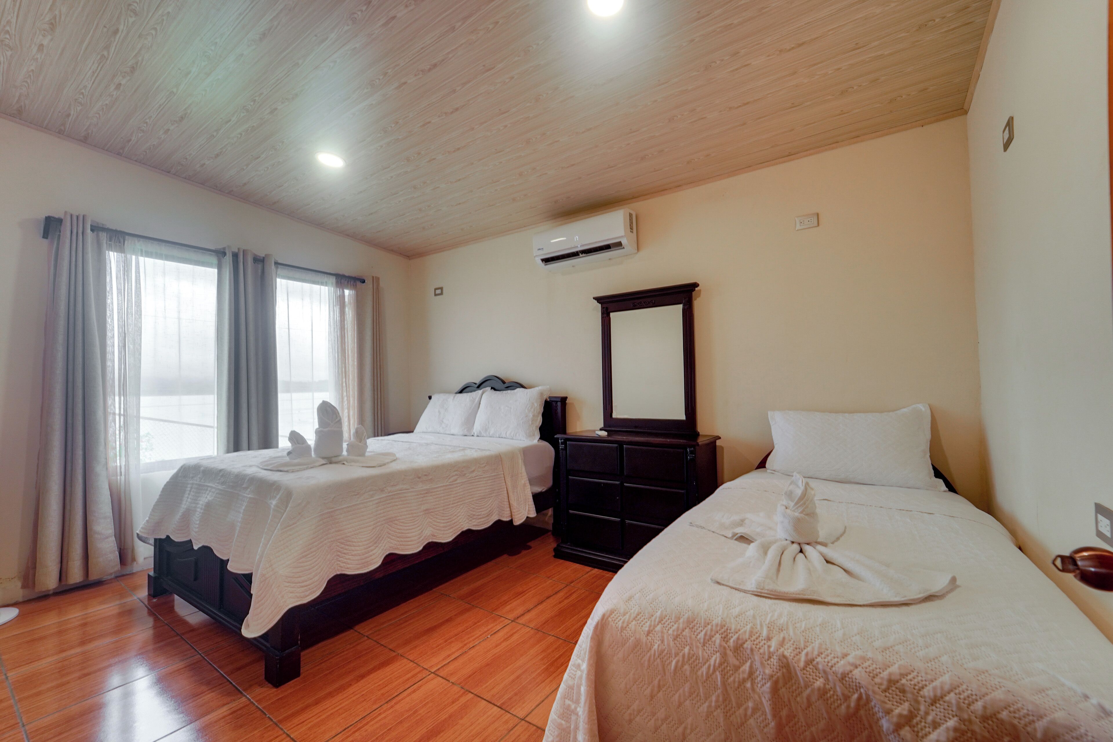 A hotel room with two beds, a wooden ceiling, an air conditioning unit, and a large window with curtains.