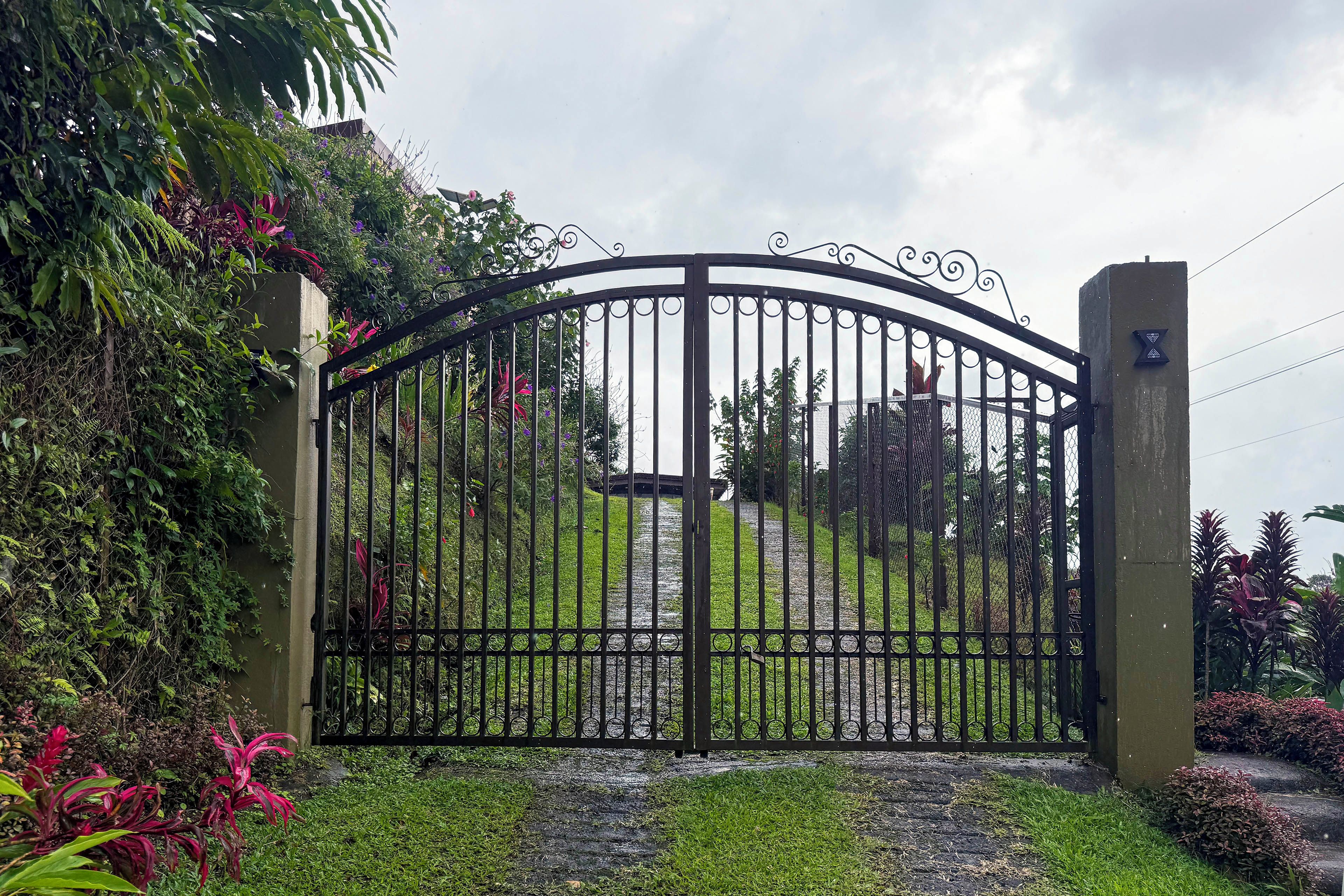A gated entrance with a metal gate, surrounded by lush greenery and a pathway leading to a building.