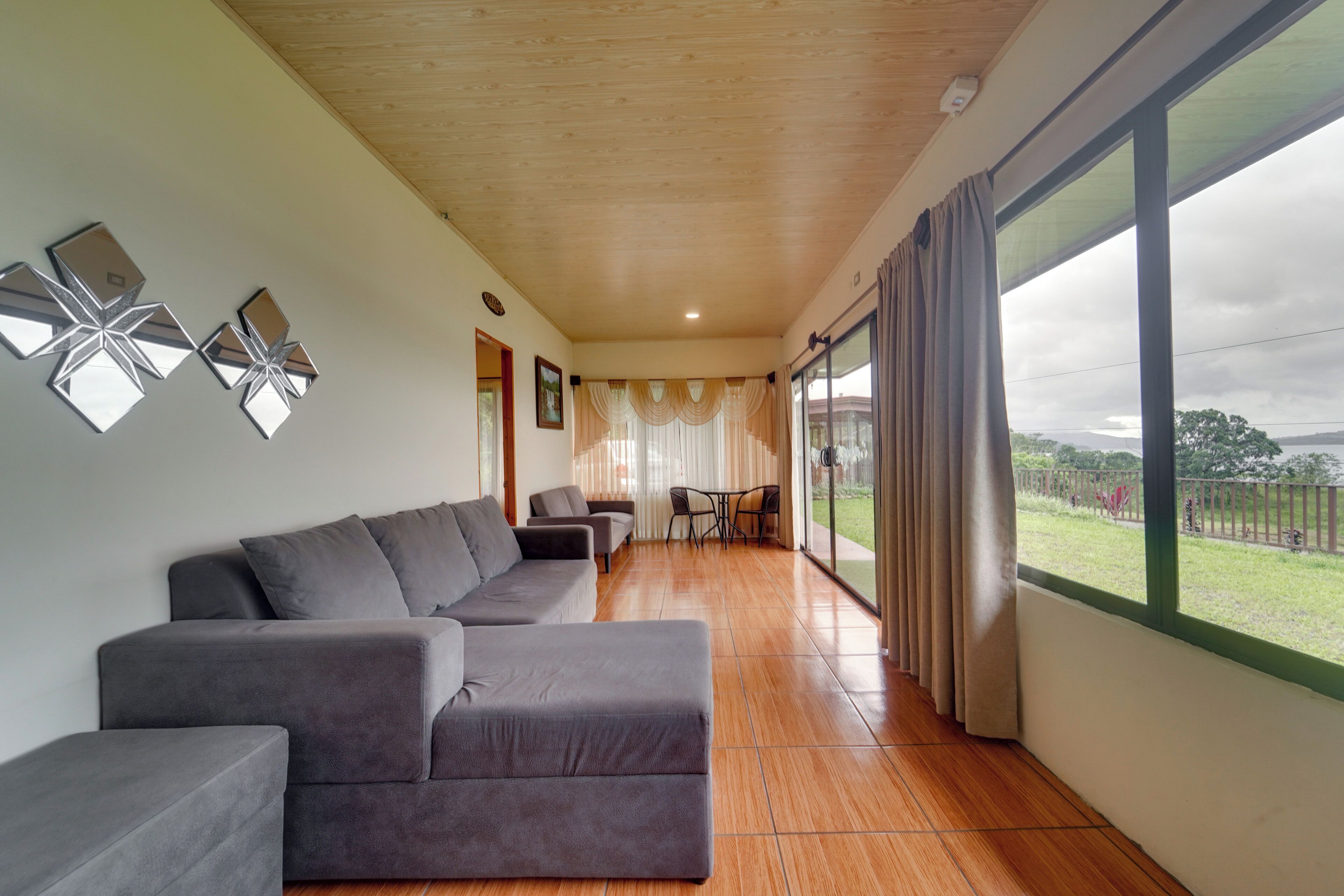 A modern living room with a grey sectional sofa, a dining area with a table and chairs, and large windows with curtains.