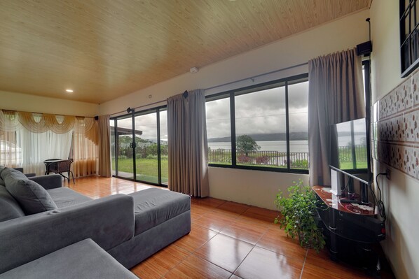 A spacious living area with a grey sofa, a dining table, and a large window offering a view of a body of water.