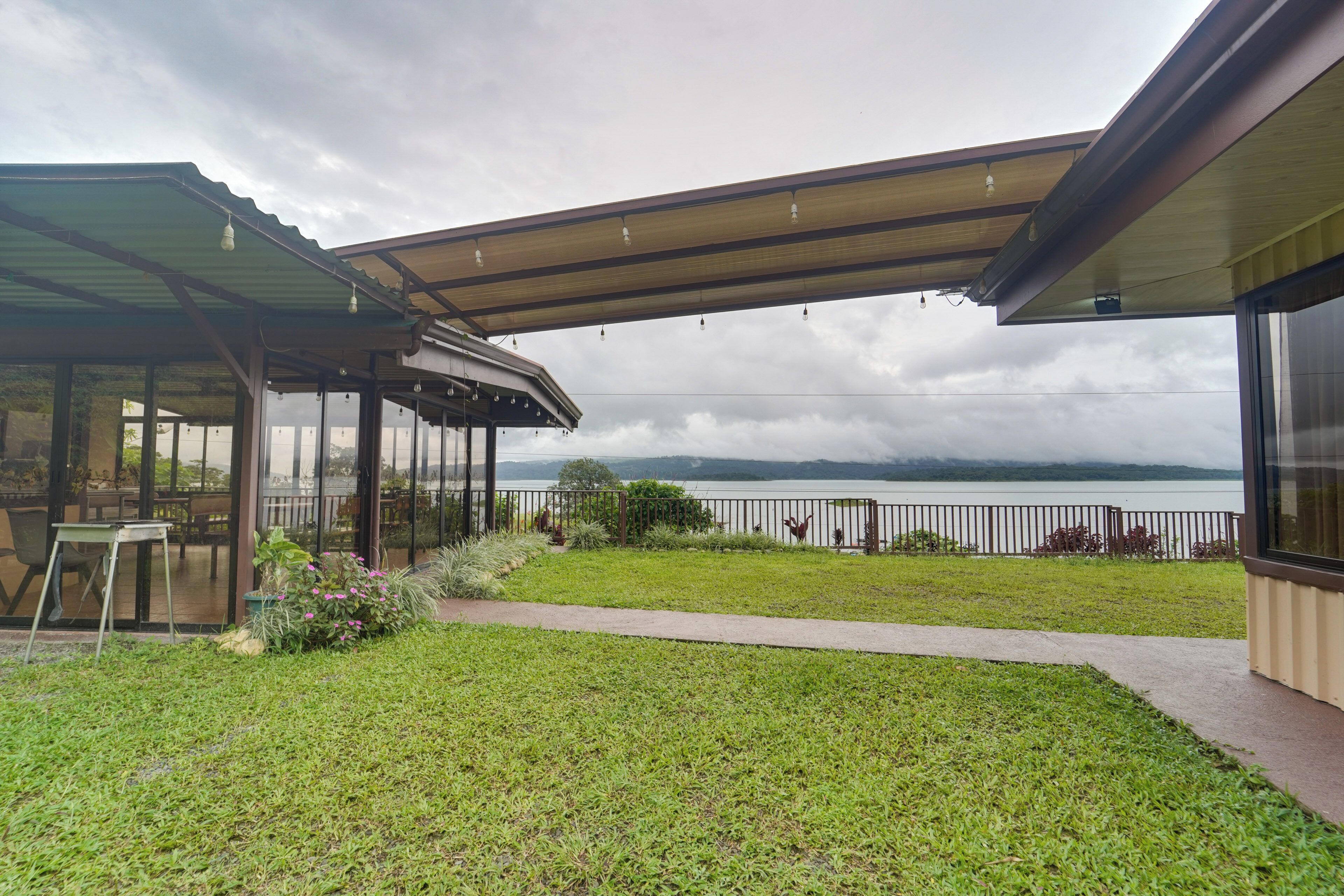 A covered outdoor area with a view of a body of water, a grassy lawn, and a seating area with tables and chairs.
