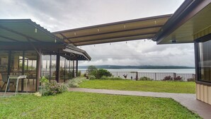 A covered outdoor area with a view of a body of water, a grassy lawn, and a seating area with tables and chairs. - Home w/ Surrounding Lake Arenal & Volcano Views! (Peñas Blancas)