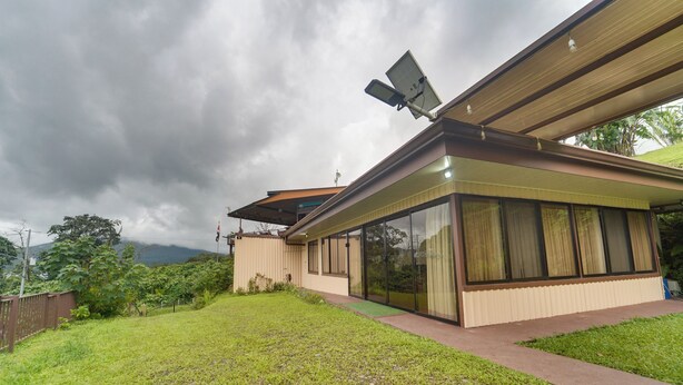 A modern house with a large glass window, a solar panel, and a grassy yard. - Home w/ Surrounding Lake Arenal & Volcano Views! (Peñas Blancas)