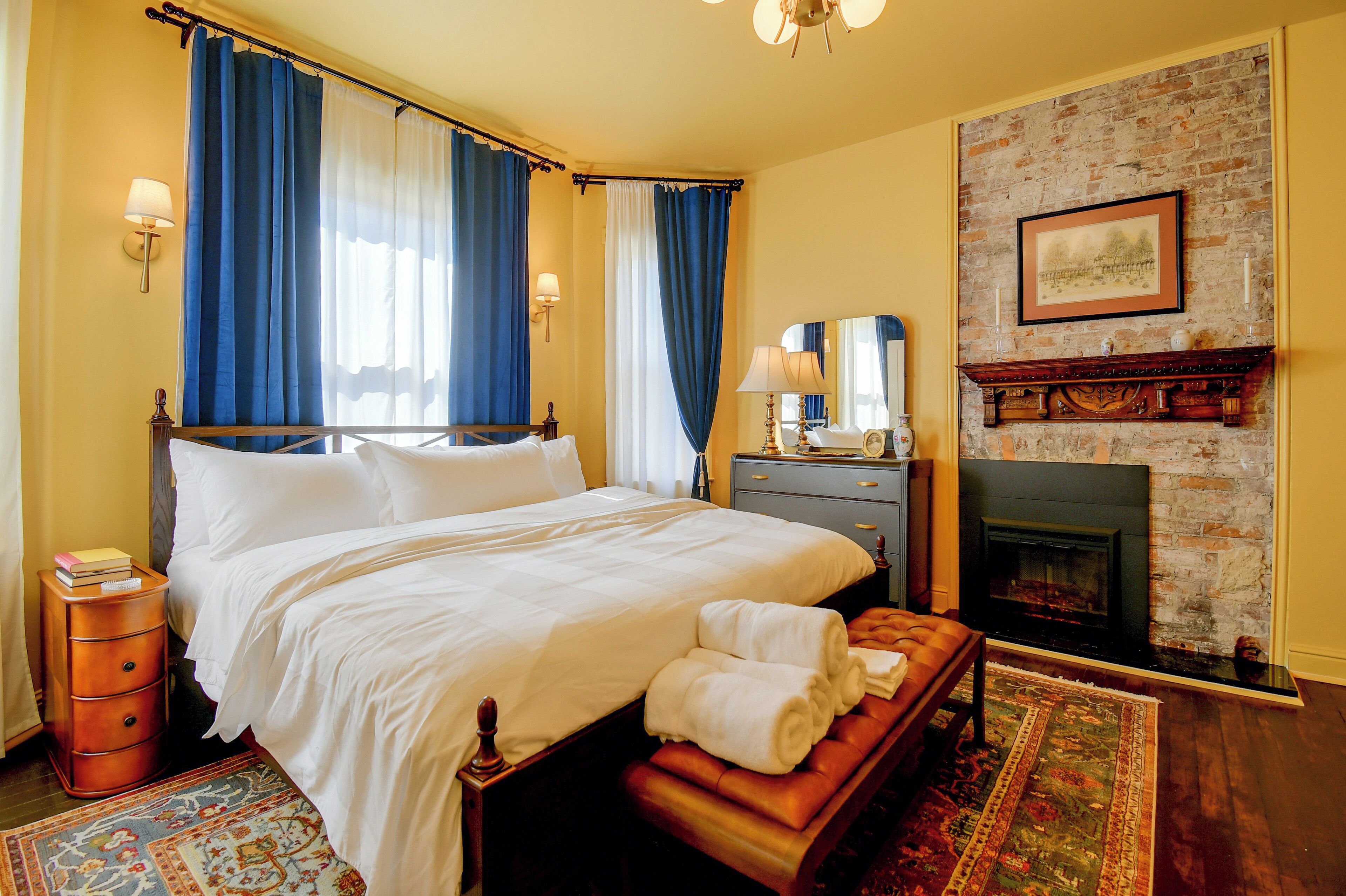 A hotel room with a large bed, a bedside table, a nightstand, a fireplace, and a rug.